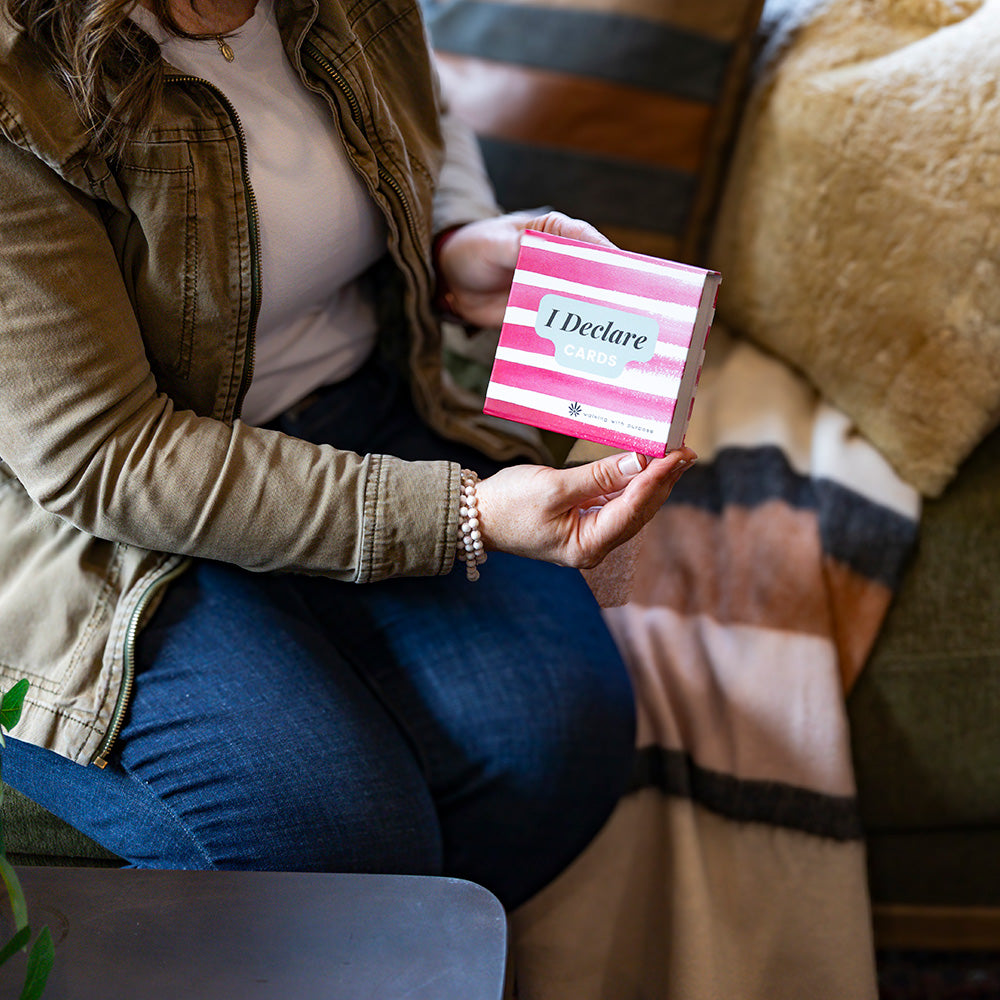 Person holding a pink and white striped card with 'I Declare Cards' text on a couch.