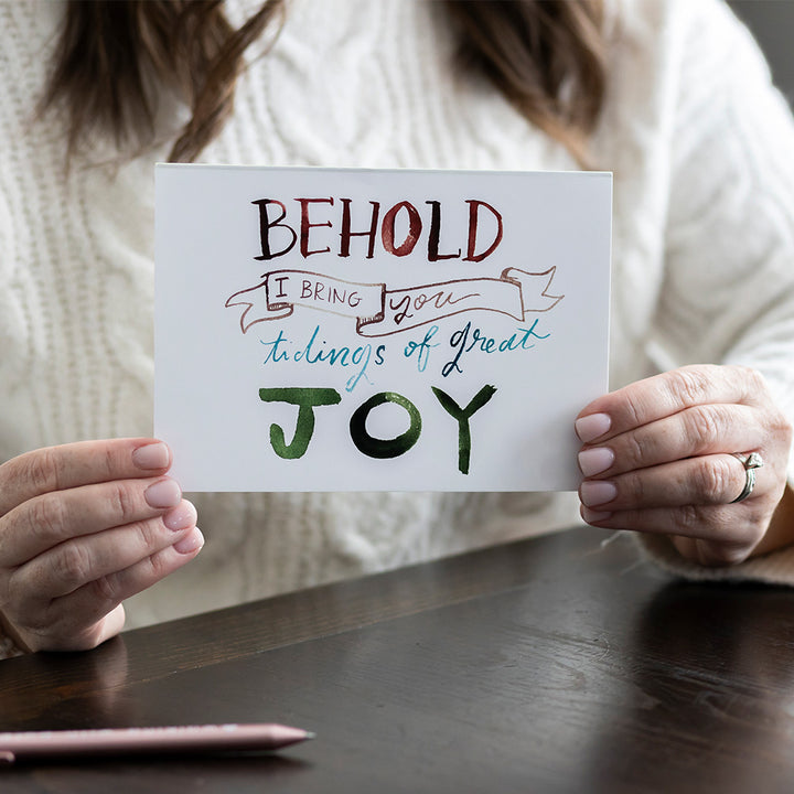 Woman holding Christmas card with decorative text "Behold I bring you tiding of great joy" on on a wooden surface with a pen