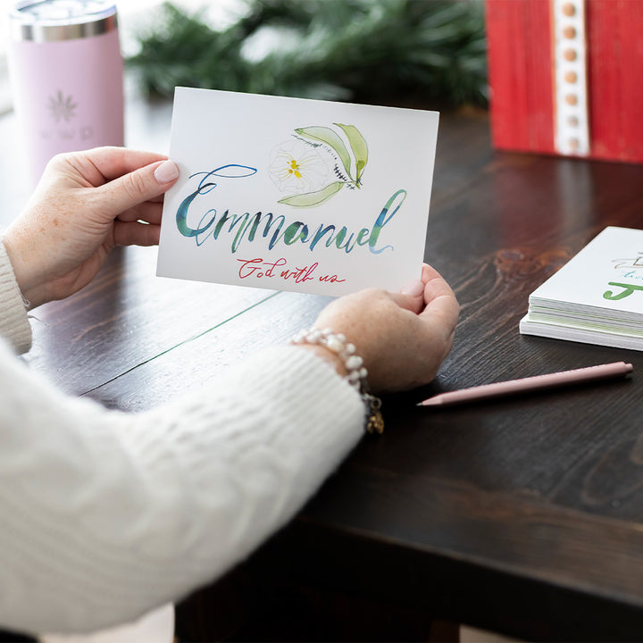 Person holding a card with 'Emmanuel God is with us' and a lily design on a wooden table.