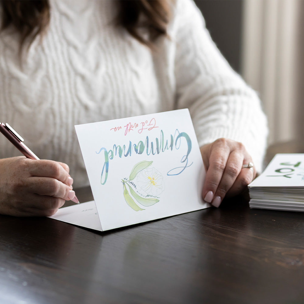 Woman writing on a Christmas card "Emmanuel God with us" with a pen, sitting at a table.