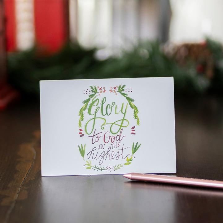 Christmas card standing up with decorative text "Glory to God in the highest" on on a wooden surface with a pen