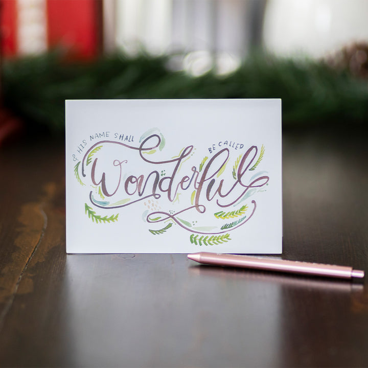 Christmas card standing up with decorative text "And His name shall be called wonderful" on on a wooden surface with a pen