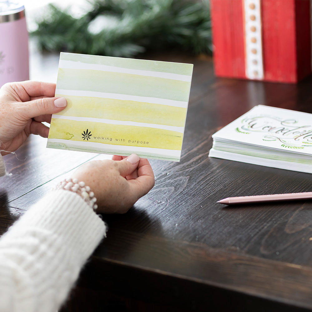 Person holding a Christmas card showing the back of the striped card on a wooden table with other cards and a pen.