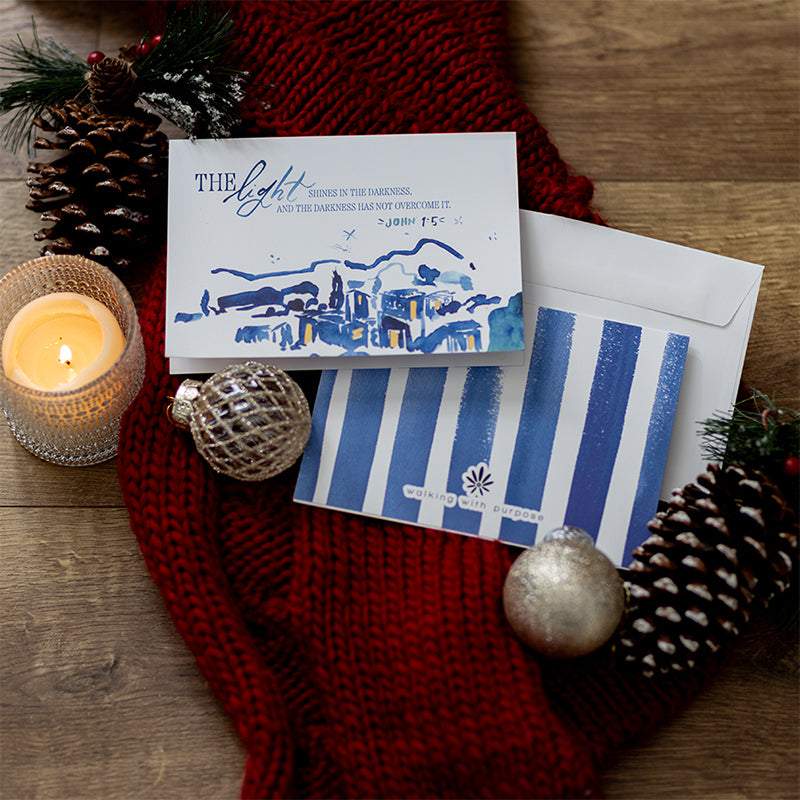 The Walking with Purpose Limited Edition Christmas Card Set features beautiful watercolor artwork in blues and yellows and the verse John 1:5: “A light shines in the darkness, and the darkness has not overcome it.” The back cover has blue stripes.