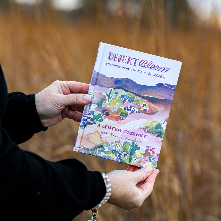 Woman holding a book titled 'Desert Bloom' with desert-themed cover against a blurred natural background.