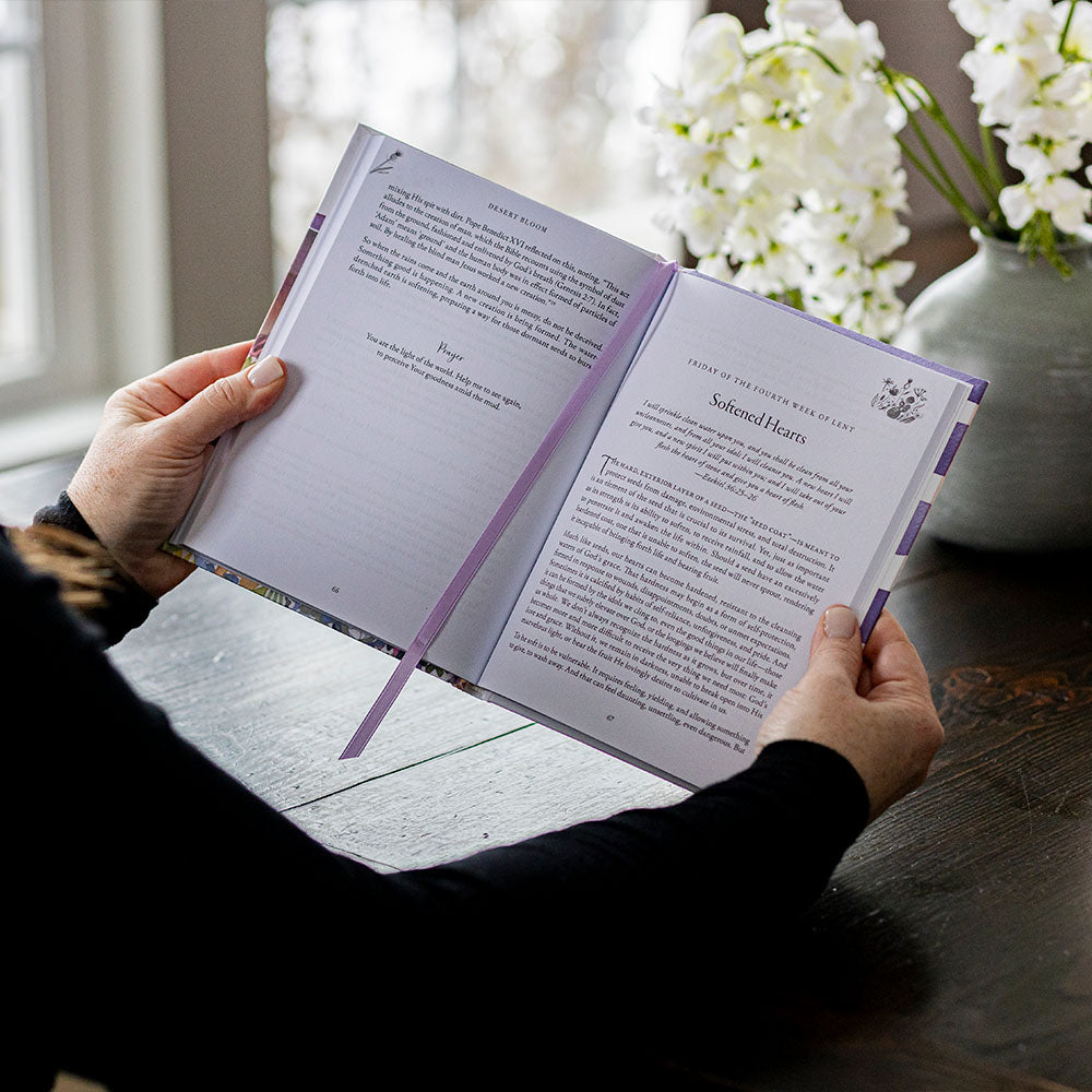 Woman holding an open "Desert Bloom" Lenten devotional with a decorative ribbon, with flowers in the background.