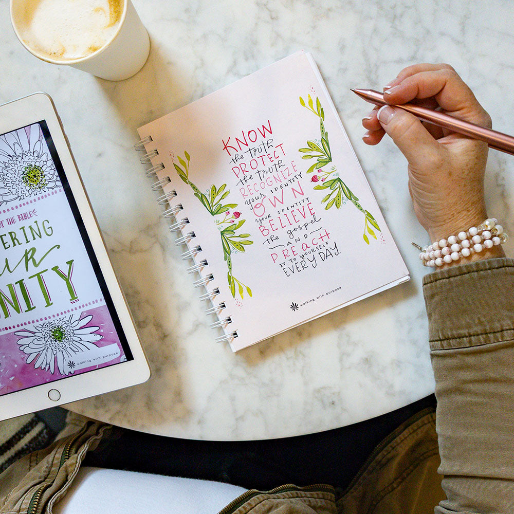 Woman holding a pen over a pink spiral-bound notebook with Fearless and Free quote,  next to a tablet displaying an e-book titled 'Discovering Our Dignity' on a marble surface
