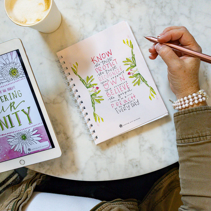 Woman holding a pen over a pink spiral-bound notebook with Fearless and Free quote,  next to a tablet displaying an e-book titled 'Discovering Our Dignity' on a marble surface