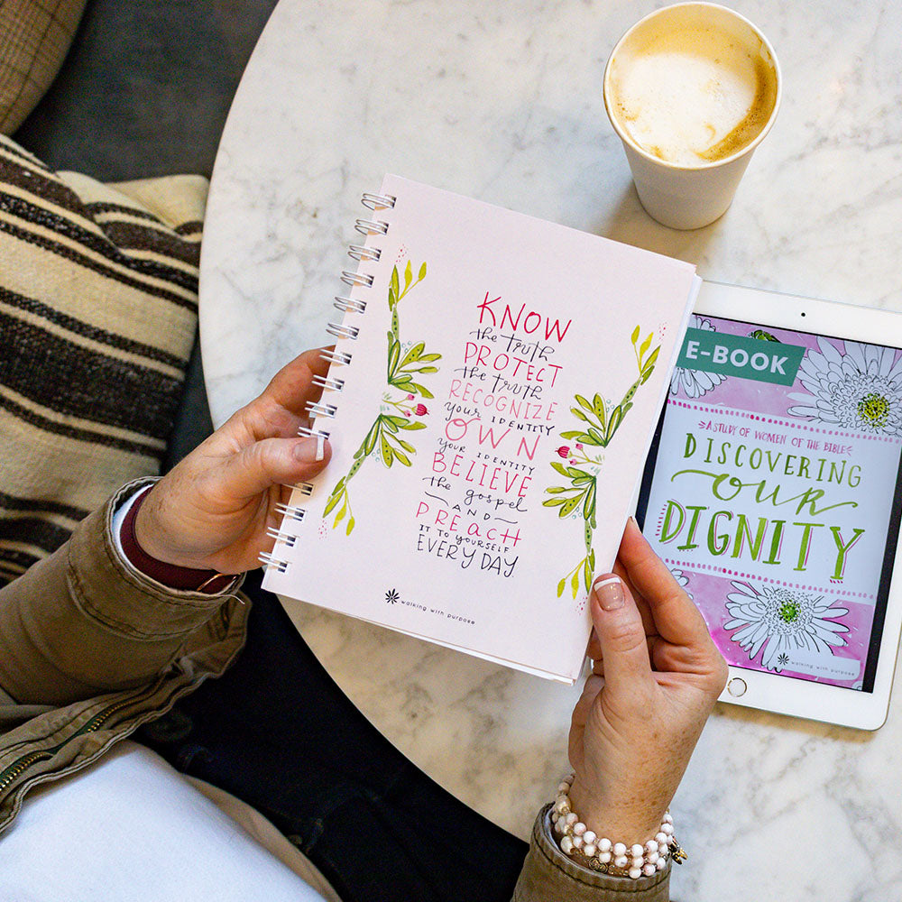 Woman holding a spiral-bound pink notebook with Fearless and Free quote next to a tablet displaying an e-book titled 'Discovering Our Dignity'.