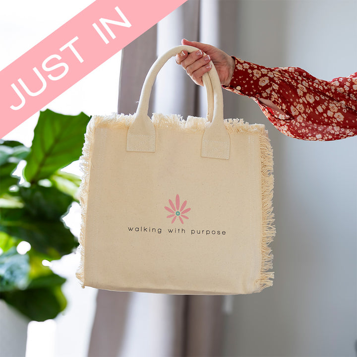 woman holding cream colored fringe tote bag with Walking with Purpose logo and Just In banner