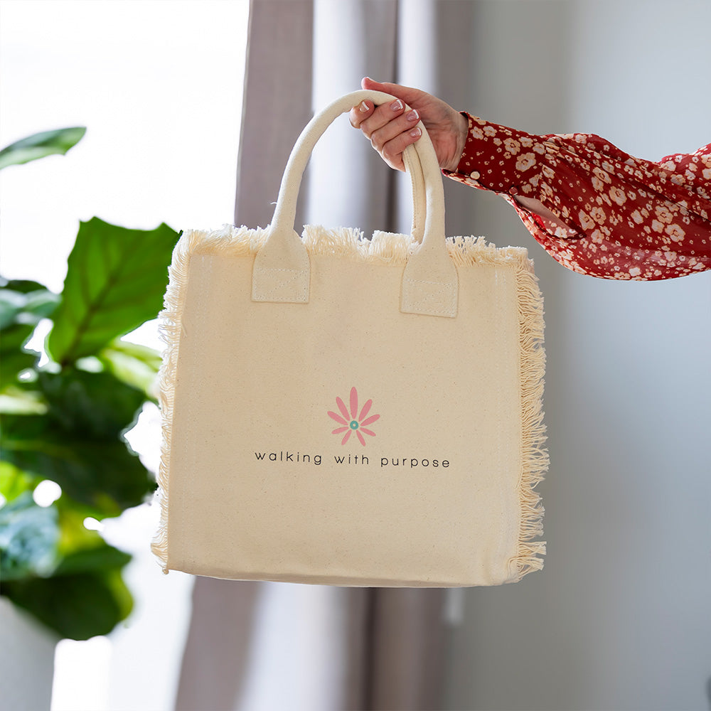 woman holding cream colored fringe tote bag with Walking with Purpose logo