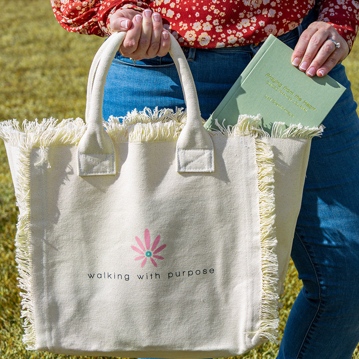 woman adding a book to cream colored fringe tote bag with Walking with Purpose logo