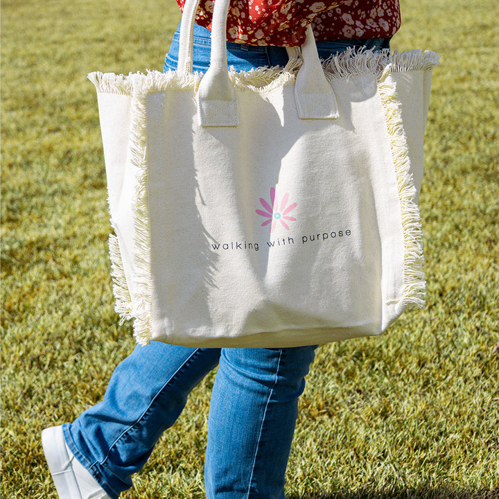 woman walking with cream colored fringe tote bag with Walking with Purpose logo