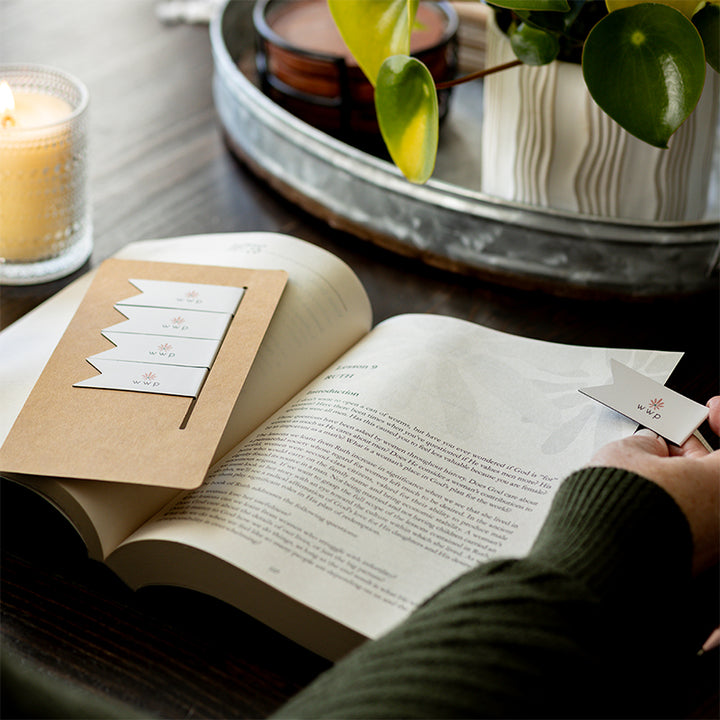 Walking with Purpose Magnetic Bookmarks designed with the signature WWP flower logo. Woman is placing one bookmark onto Bible study page. Plant and remaining bookmarks in the background.
