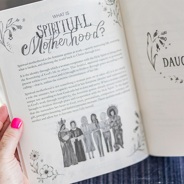 A hand with bright pink nail polish holds an open book showing printed pages about spiritual motherhood, with floral illustrations and black text on off-white paper.