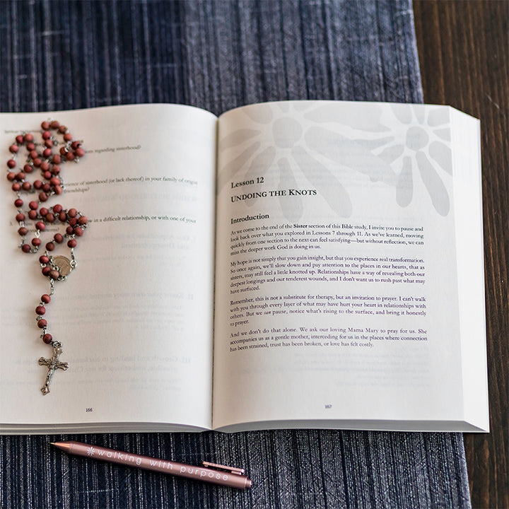 An open book, Spiritual Motherhood, spread showing printed text and a rosary draped across the left page, placed on a dark textured fabric surface with two pink pens at the bottom.