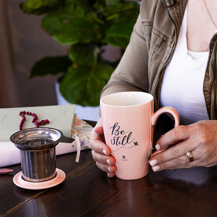 A woman holding a Light pink Be Still Tea Mug with Infuser and Lid at a table