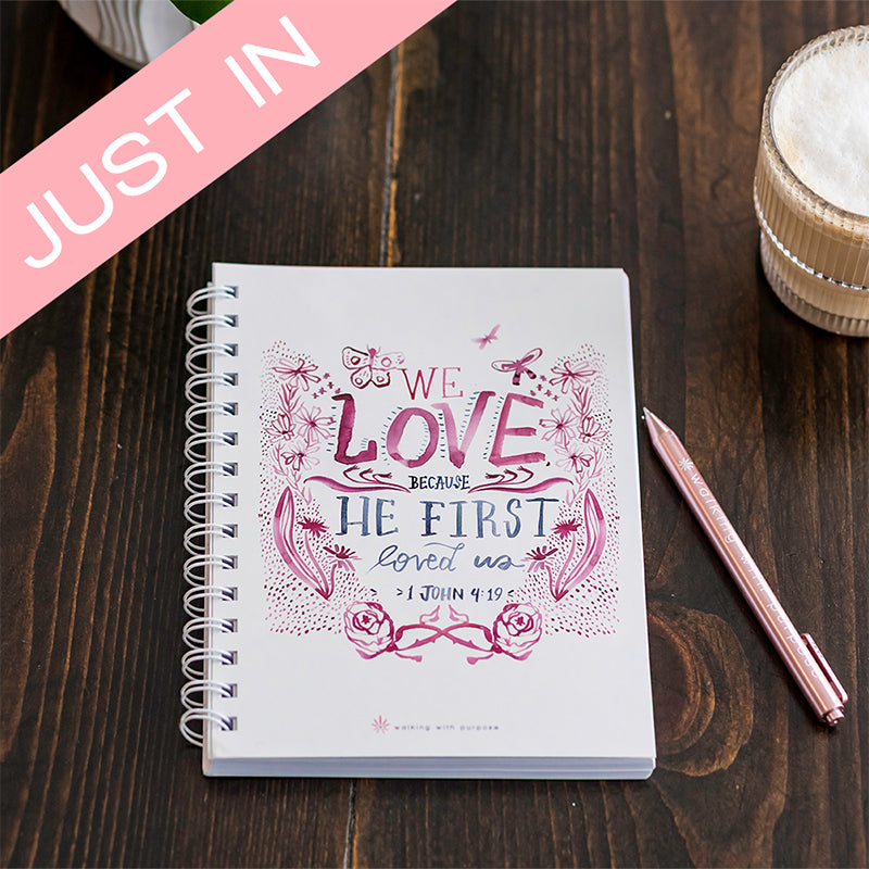 A white spiral-bound notebook with pink and blue decorative text reading 'WE LOVE BECAUSE HE FIRST loved us > John 4:19' sits on a dark wood tabletop next to a pink pen. A pink 'JUST IN' banner overlays the top-left corner.