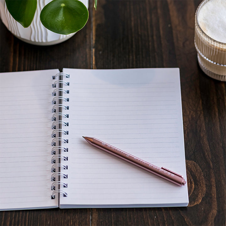A white spiral-bound notebook sits open on a dark wood tabletop next to a plant and cup of coffee with a pink pen. on top 